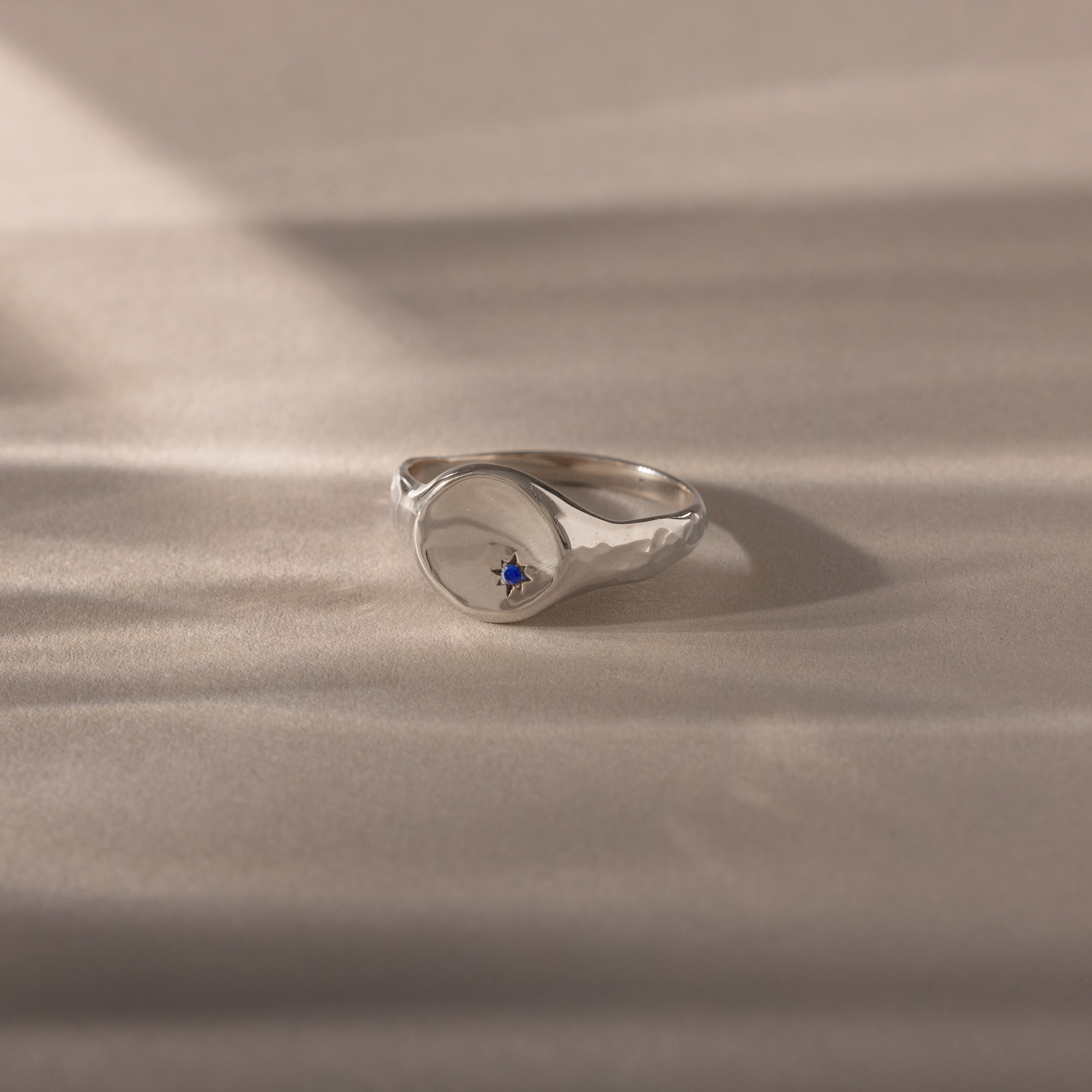 The North Star Sapphire Signet Ring, with its striking blue gemstone, rests on a smooth light beige surface with soft shadows, letting this symbolic jewelry piece truly shine.