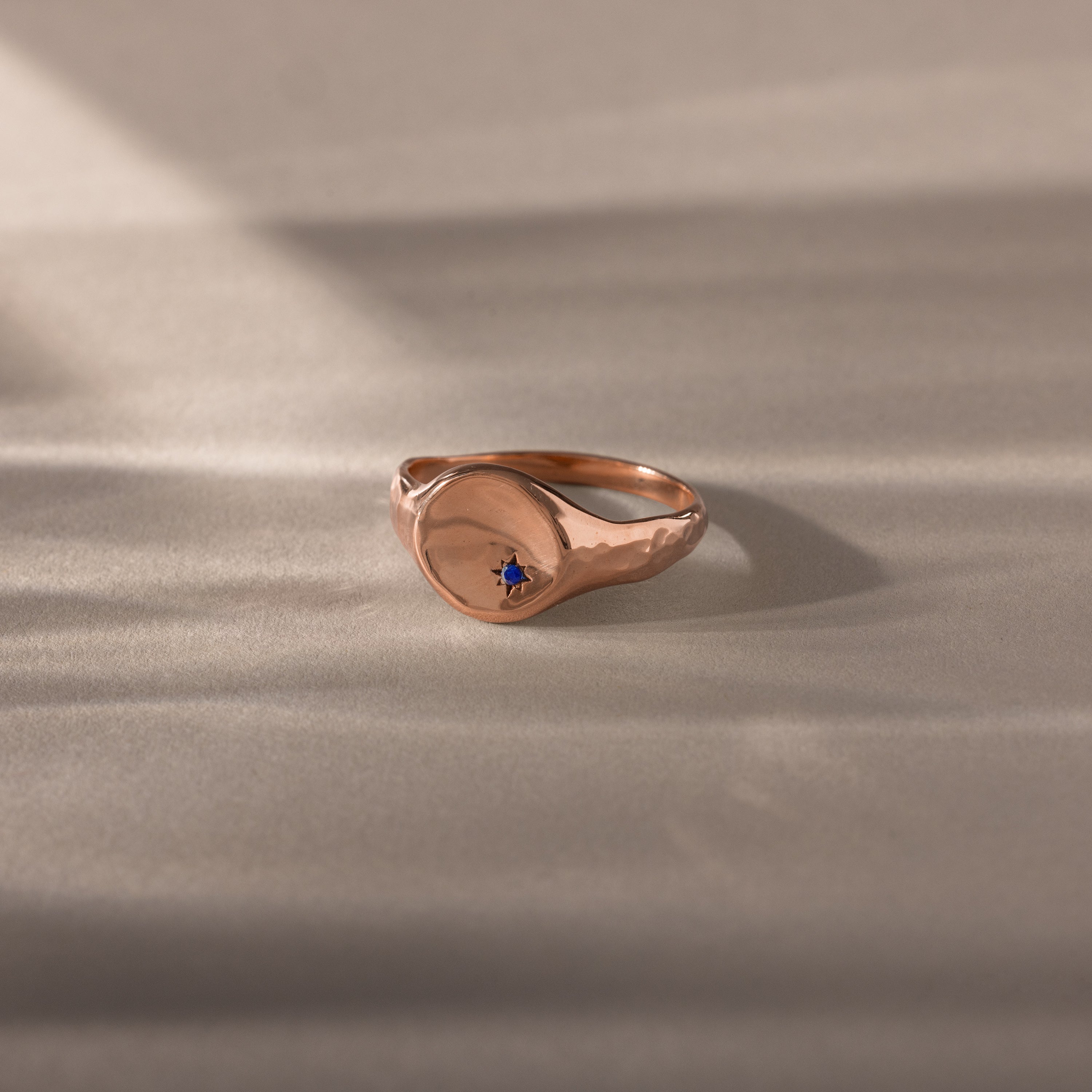 The North Star Sapphire Signet Ring features a small blue sapphire set on a smooth, round gold face, elegantly displayed on a neutral surface.
