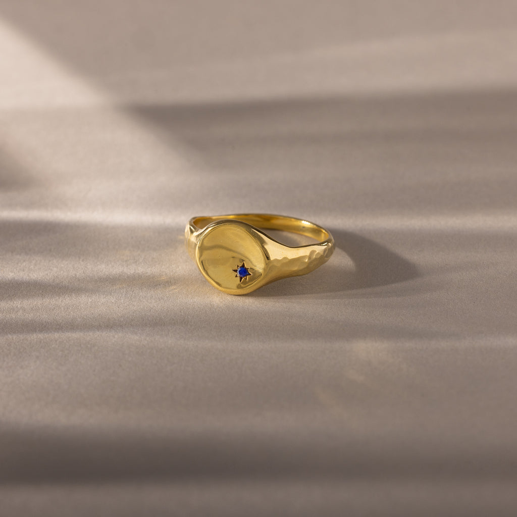 North Star Sapphire Signet Ring featuring a small blue stone, displayed on a soft beige surface with gentle shadows—a timeless piece of symbolic jewelry perfect for daily wear.