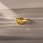 The North Star Birthstone Signet Ring, featuring an onyx center stone, is a symbolic gold ring displayed elegantly on a light, textured surface.