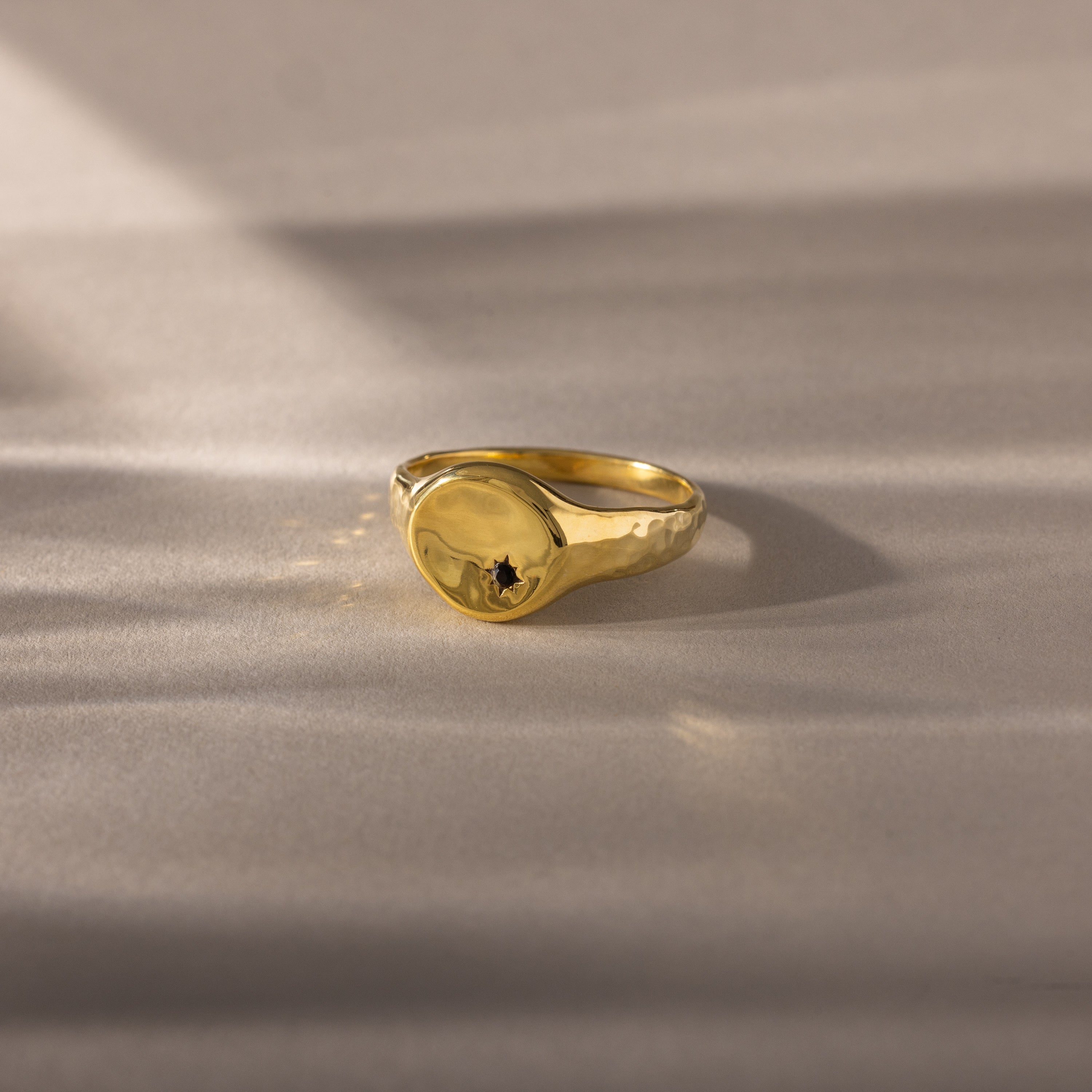 The North Star Birthstone Signet Ring, featuring an onyx center stone, is a symbolic gold ring displayed elegantly on a light, textured surface.