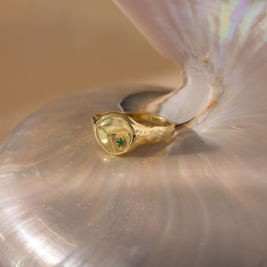 The North Star Birthstone Signet Ring, featuring a green gemstone, rests elegantly on a lustrous iridescent seashell—a refined piece of symbolic jewelry.