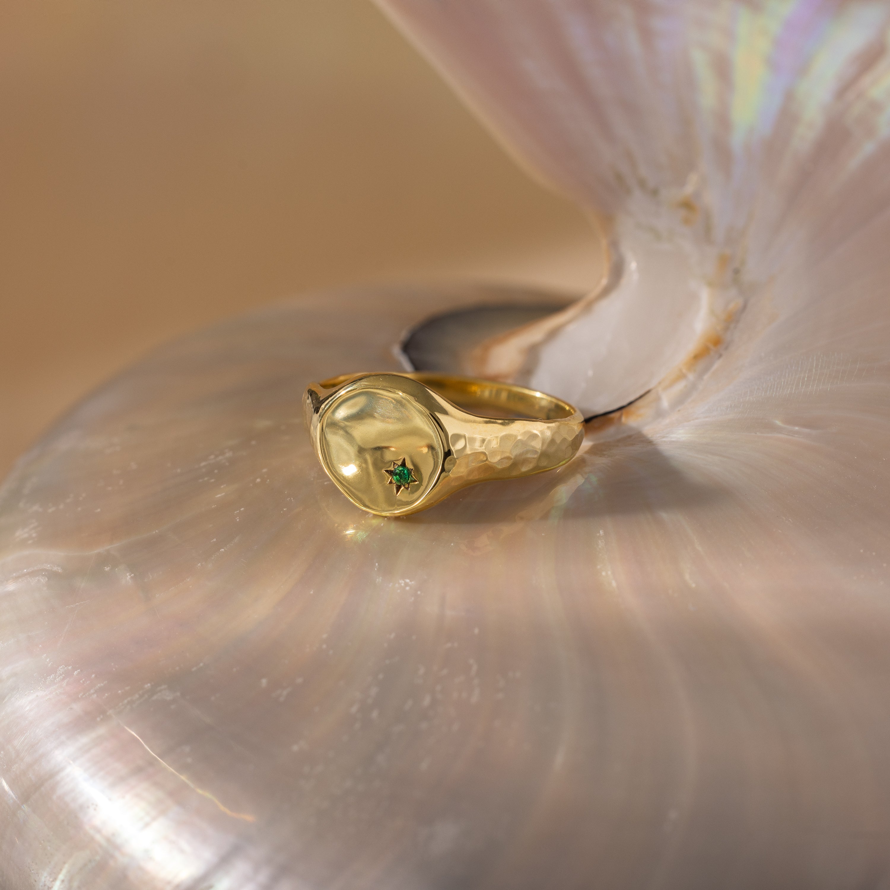 The North Star Birthstone Signet Ring, featuring a green gemstone, rests elegantly on a lustrous iridescent seashell—a refined piece of symbolic jewelry.