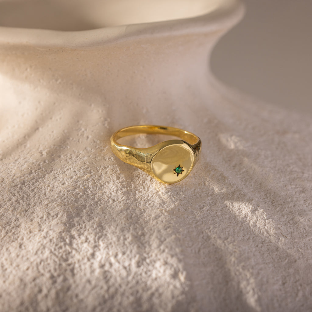 The North Star Emerald Signet Ring, featuring a green gemstone, is displayed on a textured white surface, highlighting the allure of symbolic jewelry.