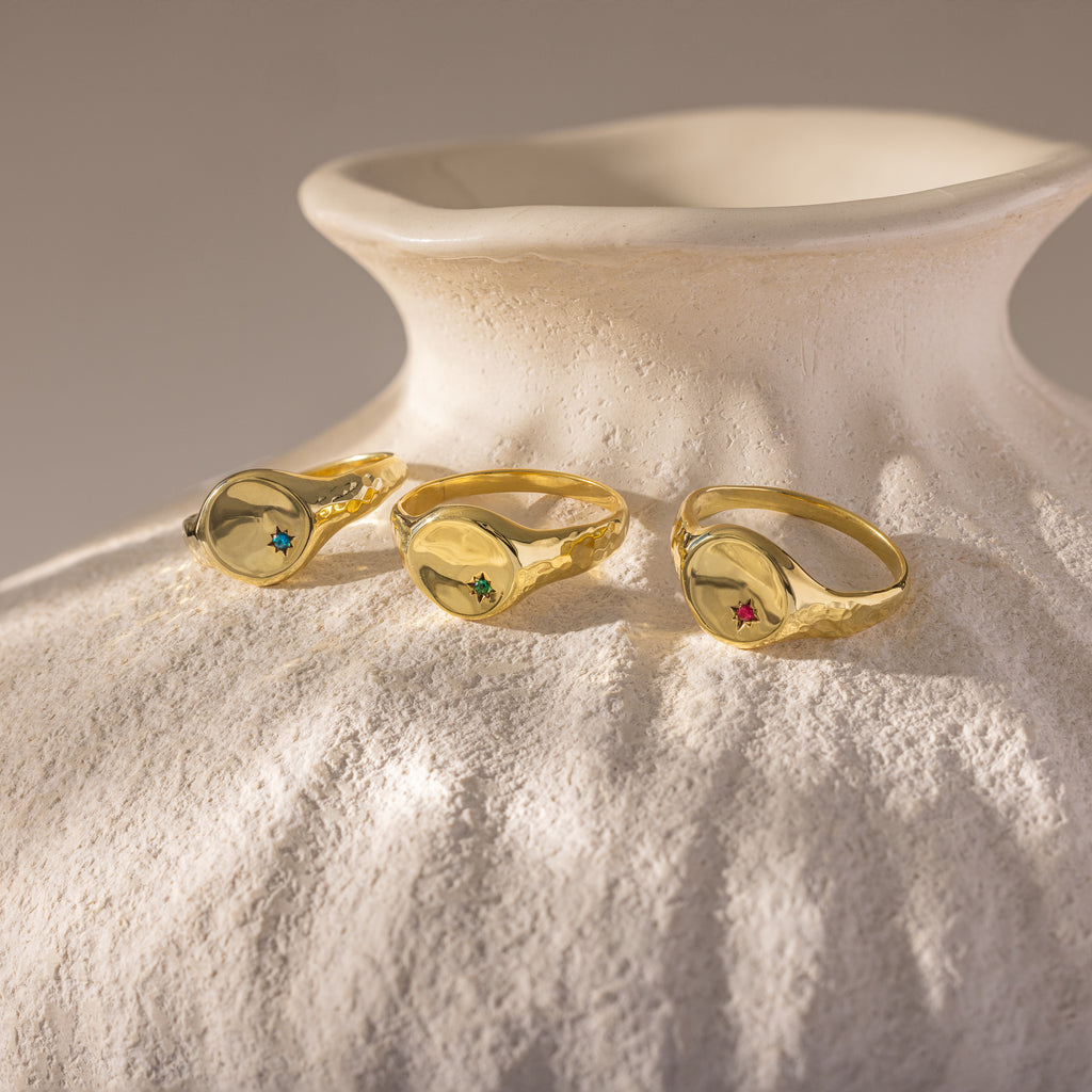 Three gold rings, including the North Star Birthstone Signet Ring with colorful gemstones, rest on a textured white ceramic vase in soft light.