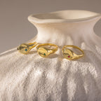 Three gold rings, including the North Star Birthstone Signet Ring with colorful gemstones, rest on a textured white ceramic vase in soft light.