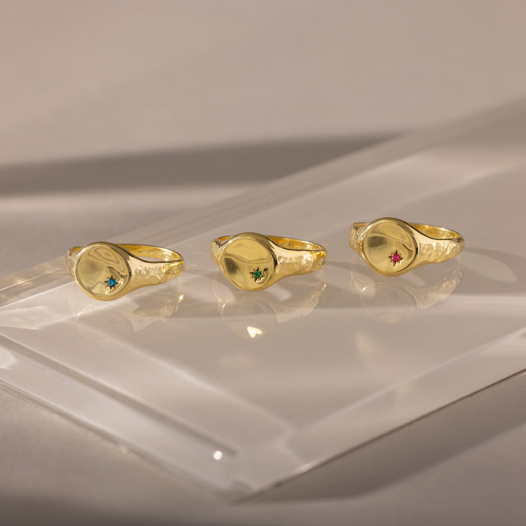 Three gold rings, including the North Star Birthstone Signet Ring with vibrant gemstones, are displayed on a reflective, translucent surface under soft lighting.