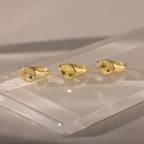 Three gold rings, including the North Star Birthstone Signet Ring with vibrant gemstones, are displayed on a reflective, translucent surface under soft lighting.