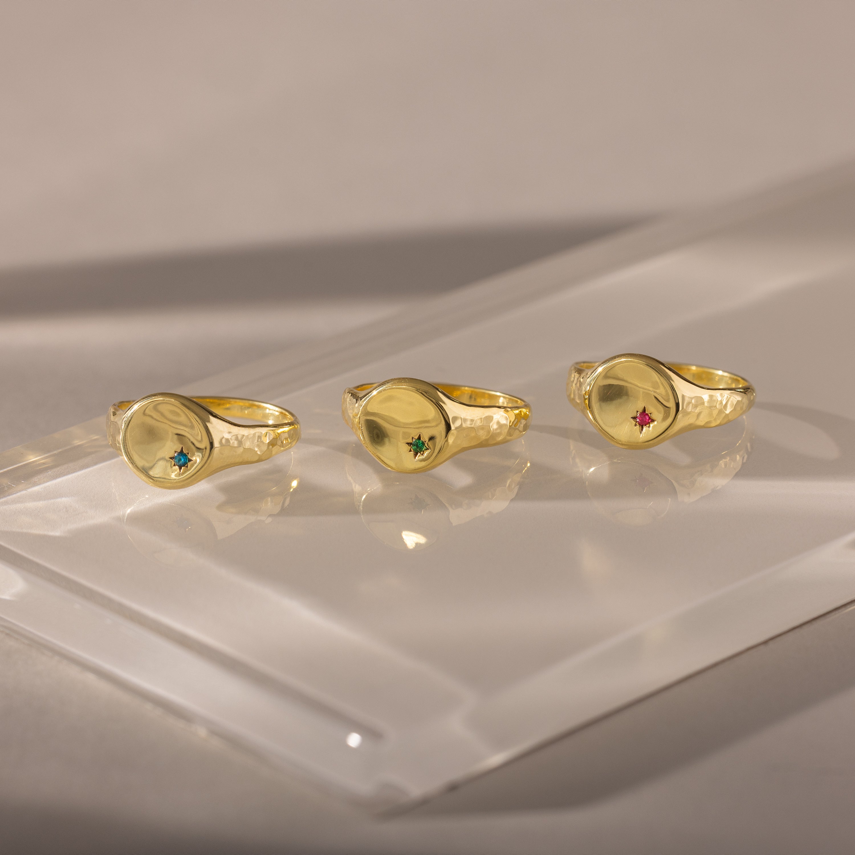 Three gold rings, including the North Star Birthstone Signet Ring with vibrant gemstones, are displayed on a reflective, translucent surface under soft lighting.