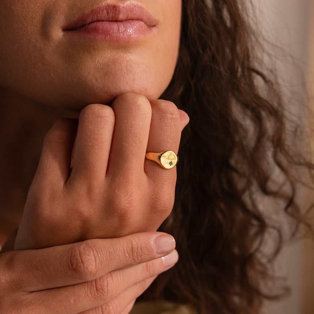 A woman with wavy hair wears a gold North Star Birthstone Signet Ring featuring a small green gemstone on her finger near her lips.