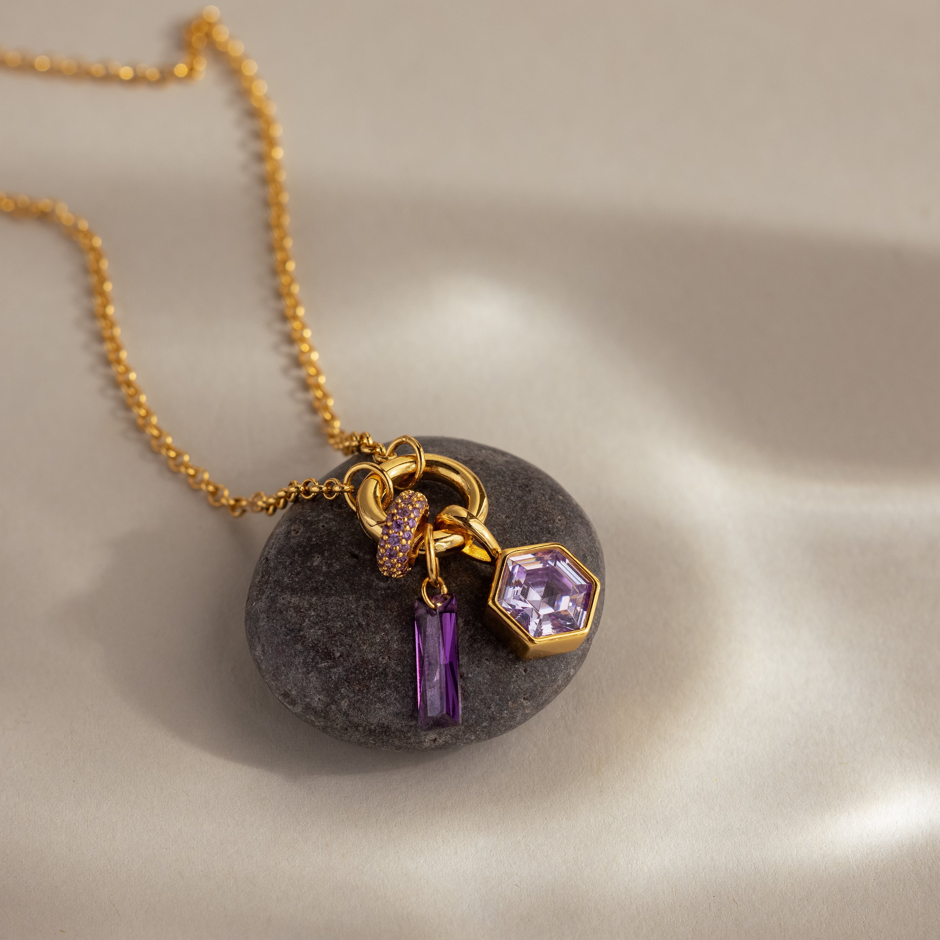 Hexagon Pink Tourmaline Charm—gold necklace with faceted stone and purple gemstone charms, showcased on a smooth gray stone; ideal for lovers of geometric jewelry.