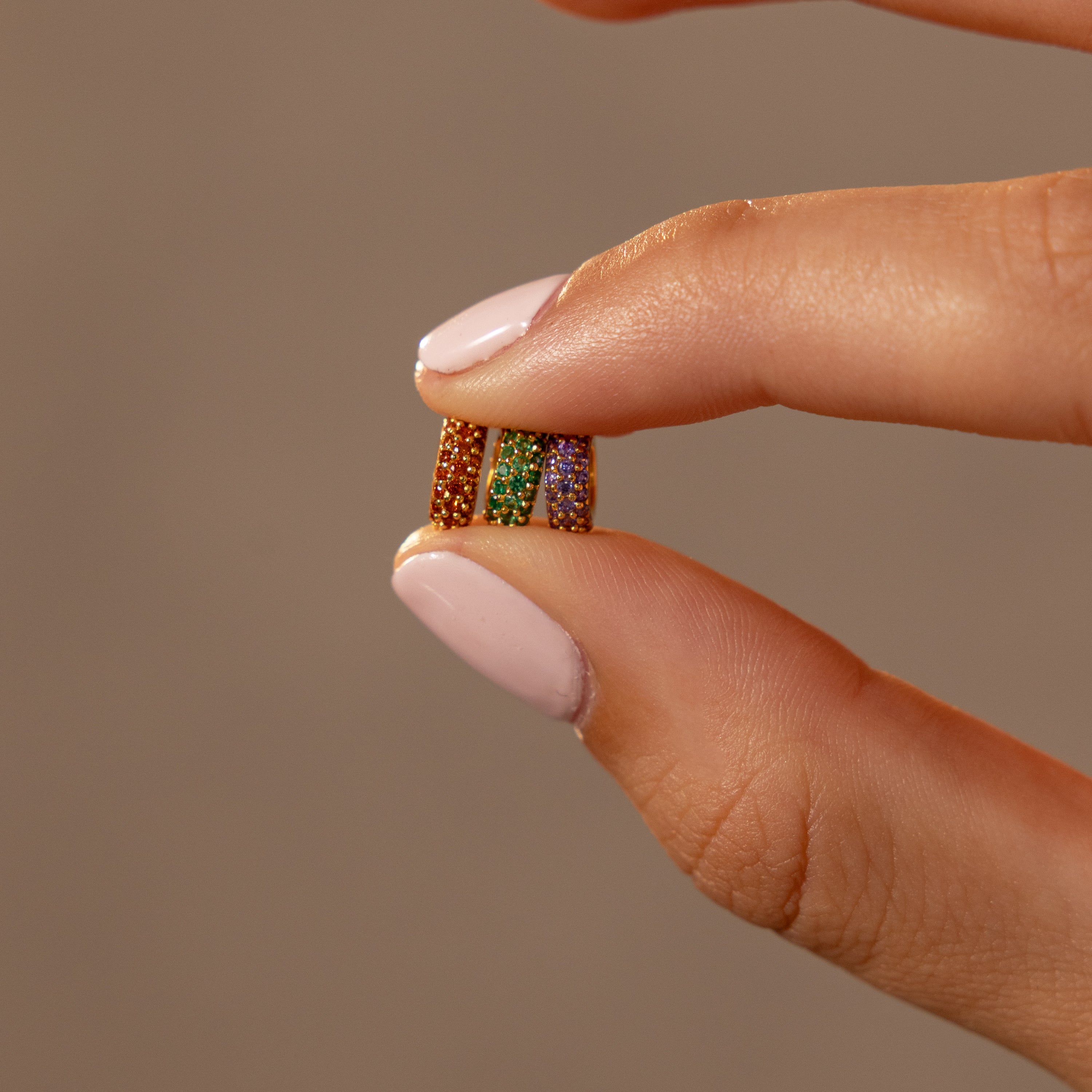 Close-up of a hand with light pink nails holding three small, colorful beaded rings between fingers, featuring a delicate Pave Emerald Charm Spacer for a personalized touch.