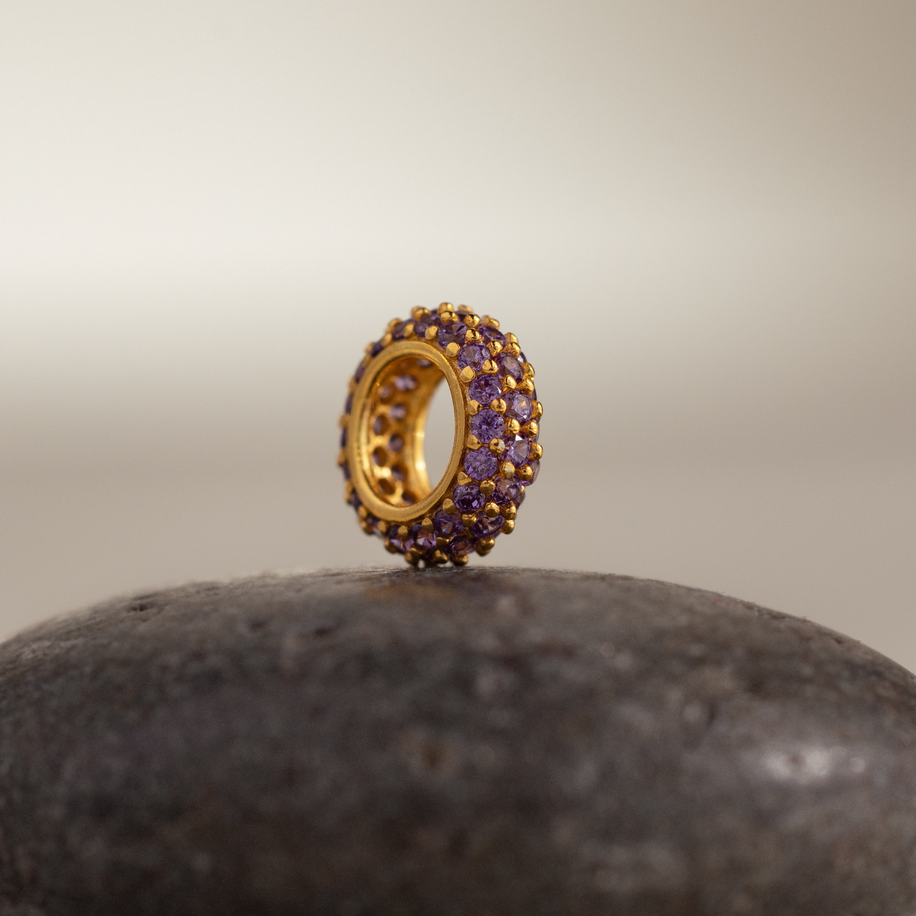 The Pave Birthstone Charm Spacer, a gold circular piece with purple gemstones, sits upright on a smooth, dark stone surface.