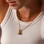 A woman in a white tank top wears layered gold necklaces with small pendants, including the Hexagon Aquamarine Charm—an elegant and meaningful gift.