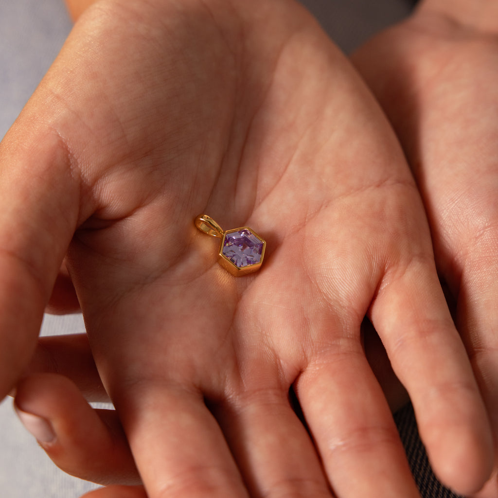 A person holds a Hexagon Alexandrite Charm with a faceted purple gemstone in their open palm.