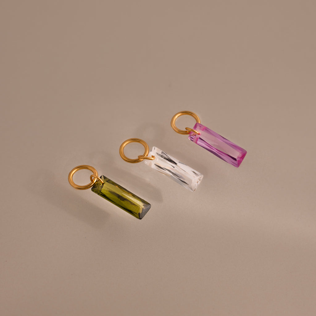 Three gold rings with Skinny Baguette Birthstone Charms—featuring green, clear, and pink rectangular crystals—are arranged on a neutral background.
