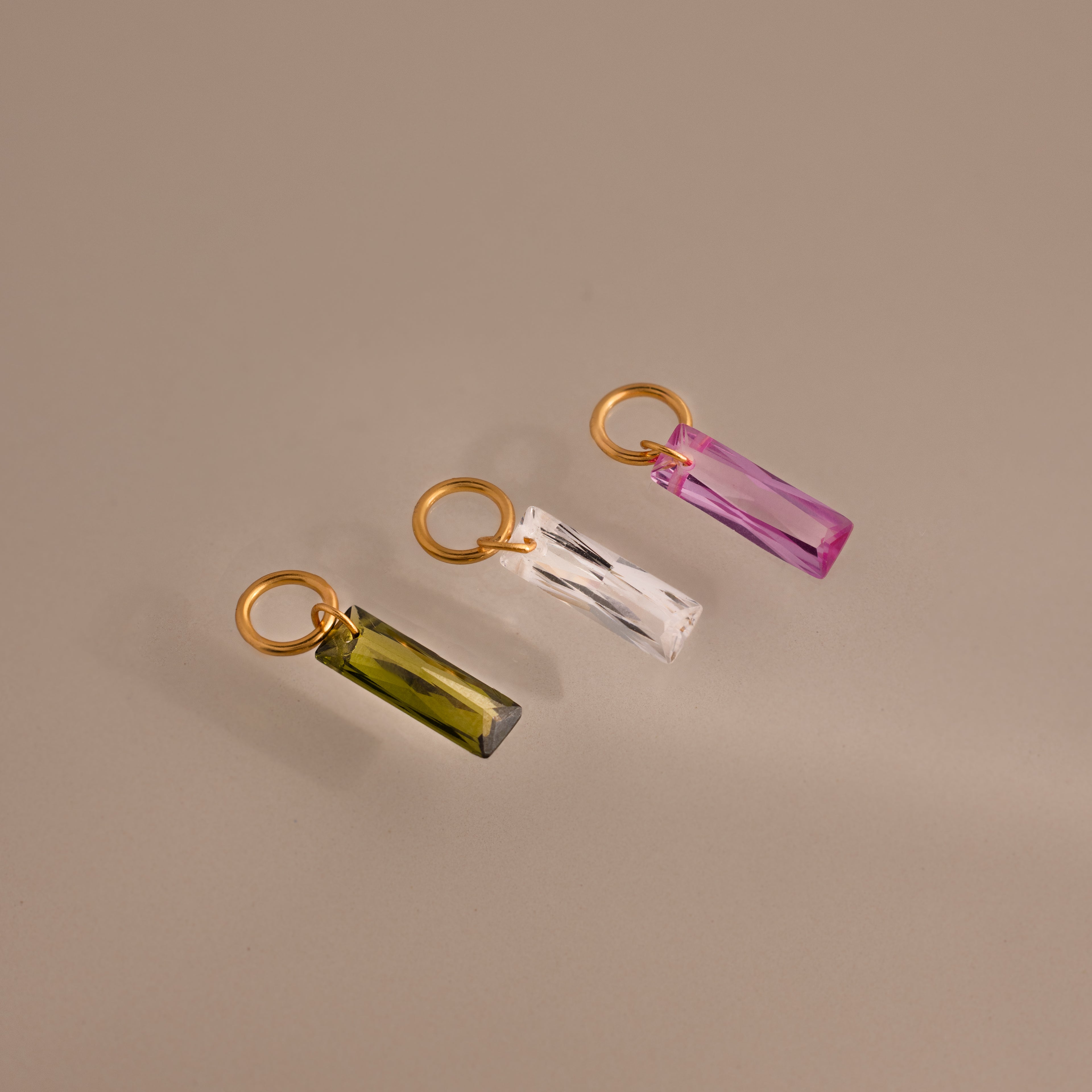 Three gold rings with Skinny Baguette Birthstone Charms—featuring green, clear, and pink rectangular crystals—are arranged on a neutral background.