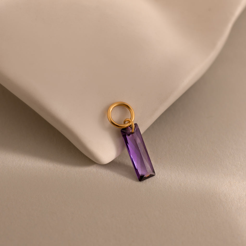 The Skinny Baguette Birthstone Charm with a rectangular purple stone rests on a soft beige fabric surface.