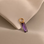 The Skinny Baguette Birthstone Charm with a rectangular purple stone rests on a soft beige fabric surface.