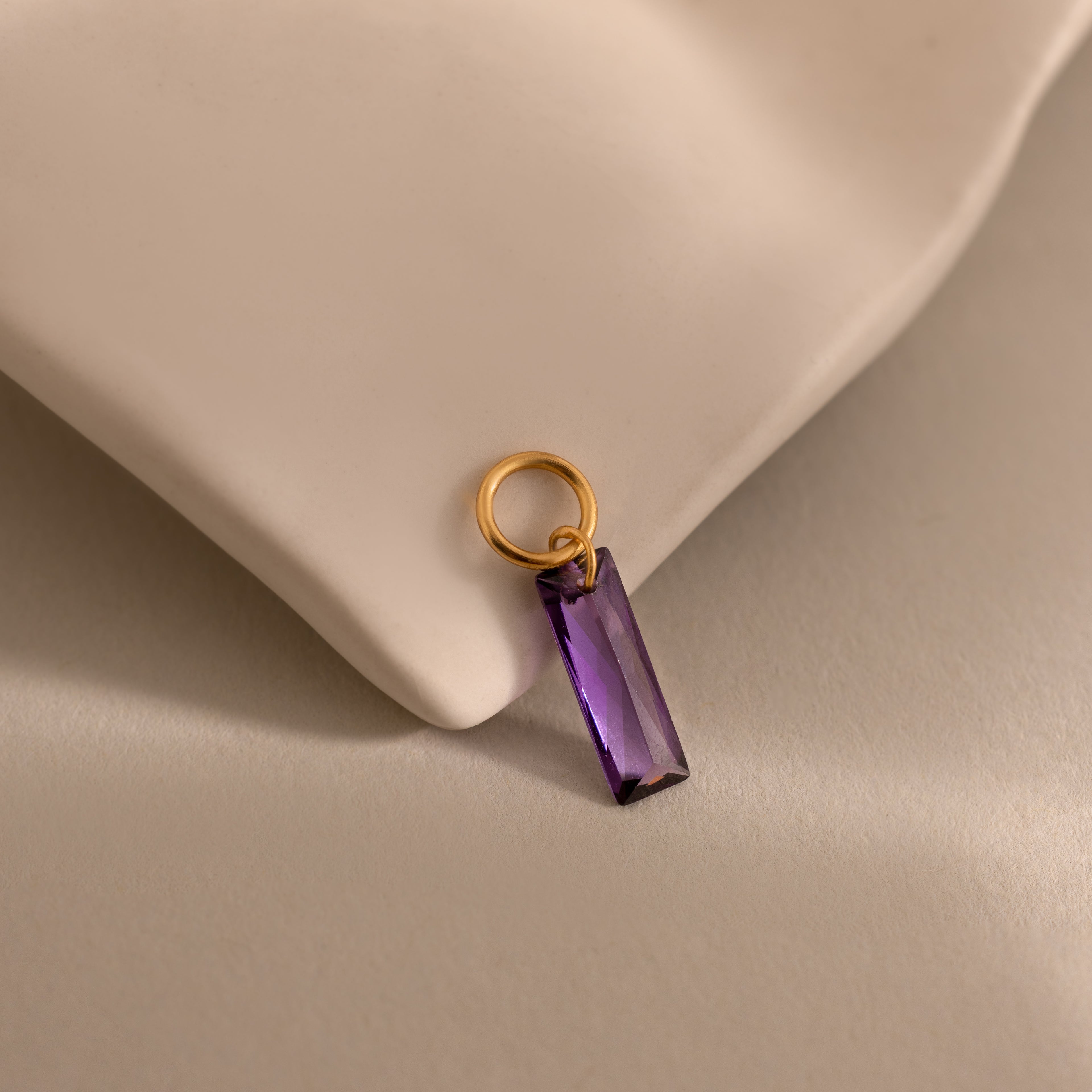 The Skinny Baguette Birthstone Charm with a rectangular purple stone rests on a soft beige fabric surface.