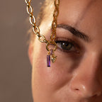 A gold chain with the Skinny Baguette Birthstone Charm in purple and gold hangs close to a womans eye.