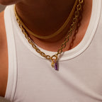A person wears layered gold necklaces with purple and gold charms, including the Skinny Baguette Birthstone Charm, over a white ribbed tank top.
