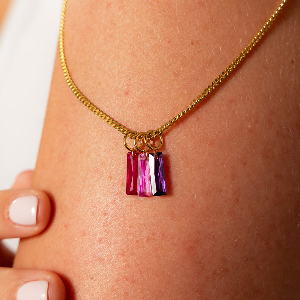 The Skinny Baguette Alexandrite Charm is a gold necklace with three rectangular pink and purple baguette-cut stones, each dangling from a delicate gold loop and worn around the neck.