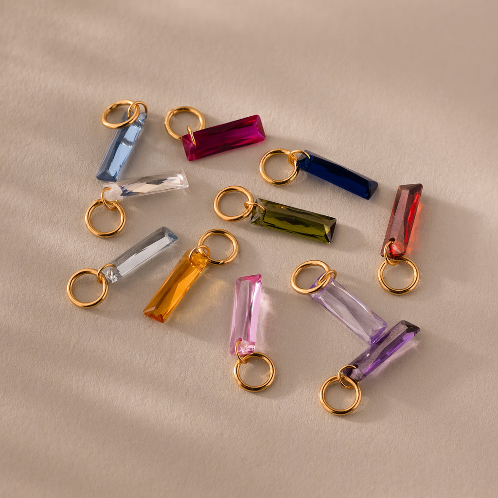 Colorful rectangular Skinny Baguette Birthstone Charms with gold rings are scattered on a beige surface.