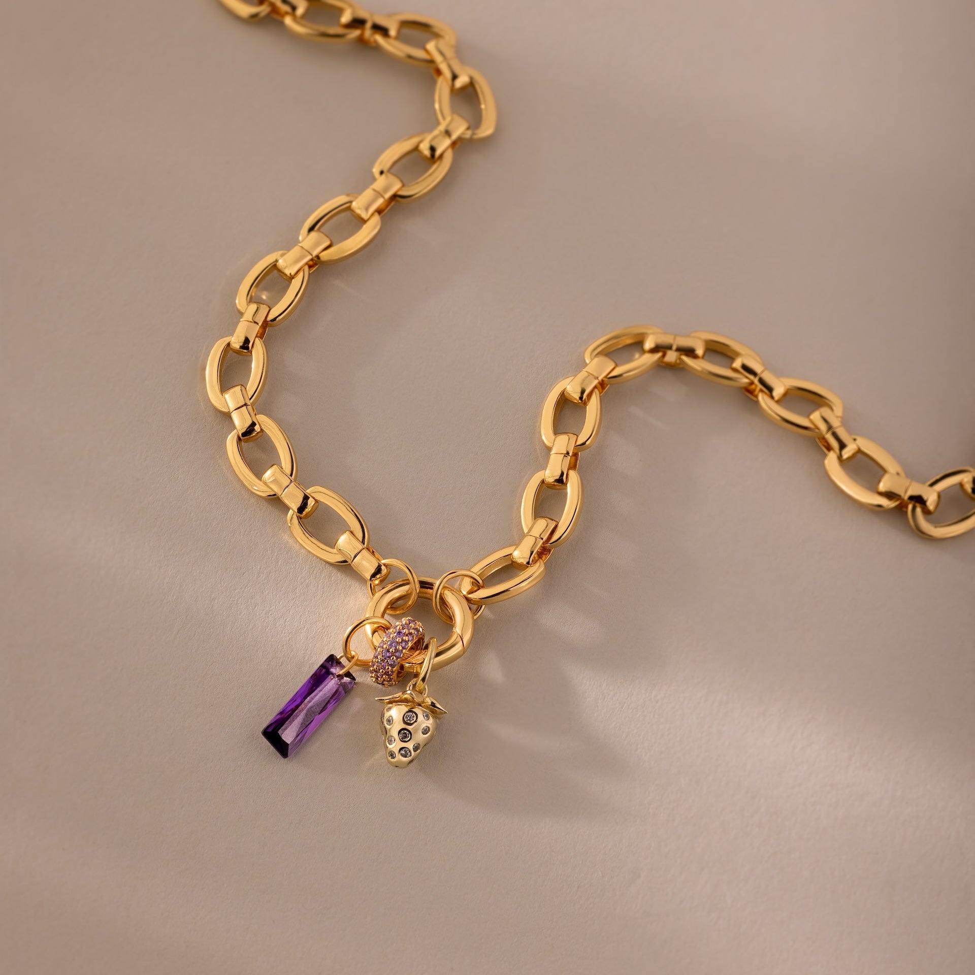 Skinny Baguette Birthstone Charm: Gold chain necklace with a gem-studded strawberry, baguette-cut birthstone, purple crystal pendant, and gold charm, shown on a beige surface.