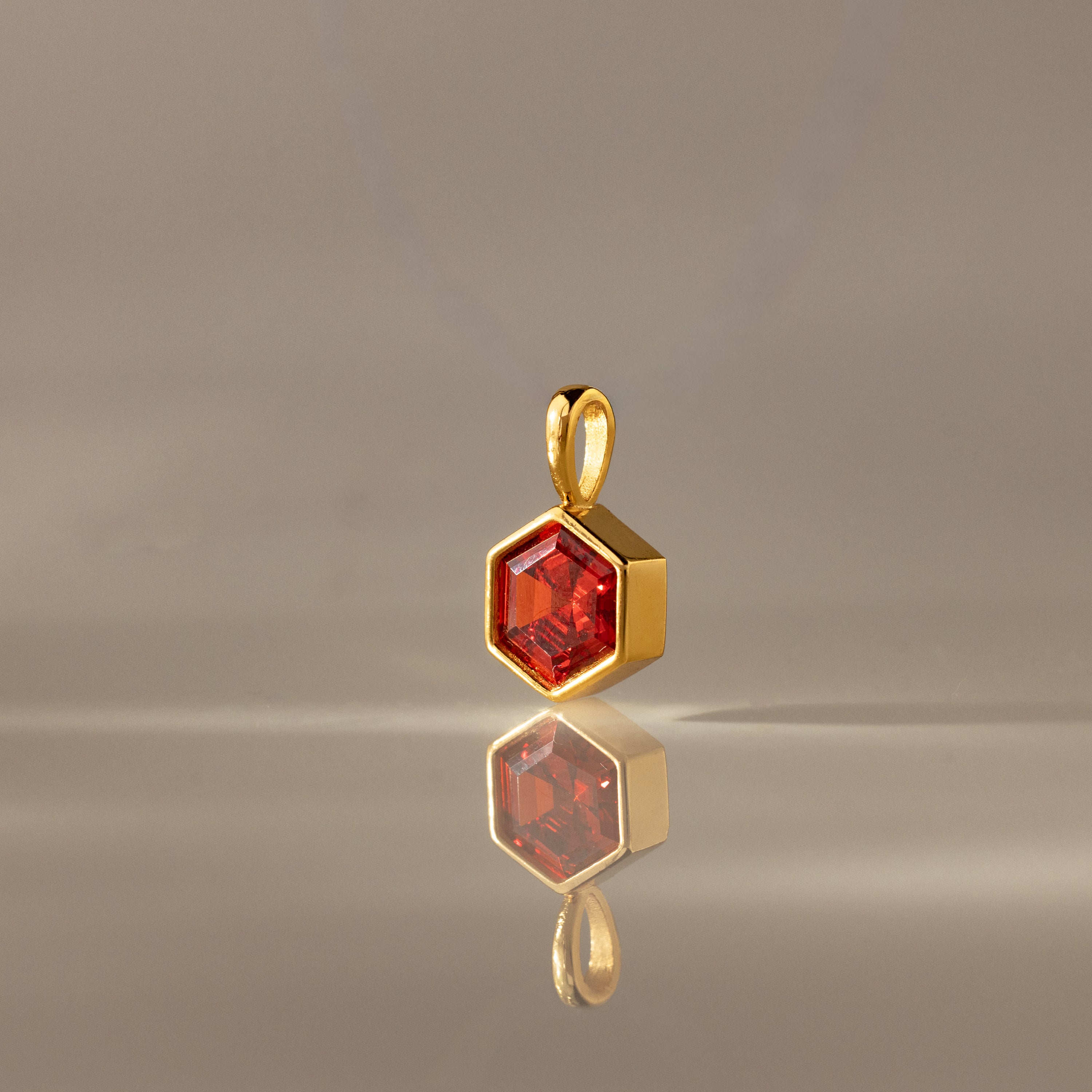 The Hexagon Garnet Charm is a gold pendant with a faceted hexagonal red gem, showcased on a smooth neutral surface—an eye-catching piece of geometric jewelry.