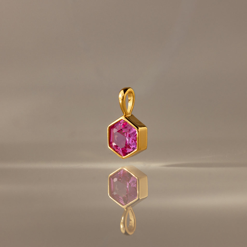 The Hexagon Pink Tourmaline Charm features a gold hexagonal pendant with a faceted pink stone, elegantly displayed on a smooth, neutral surface—a modern geometric jewelry piece.