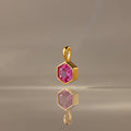 The Hexagon Pink Tourmaline Charm features a gold hexagonal pendant with a faceted pink stone, elegantly displayed on a smooth, neutral surface—a modern geometric jewelry piece.