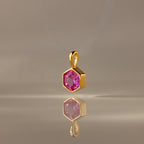 The Hexagon Pink Tourmaline Charm features a gold hexagonal pendant with a faceted pink stone, elegantly displayed on a smooth, neutral surface—a modern geometric jewelry piece.