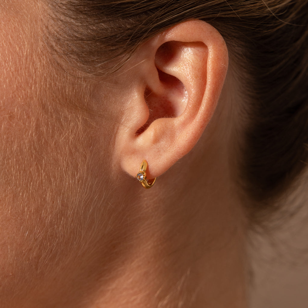 Close-up of a womans ear wearing Ribbed Aquamarine Huggies, elegant gold earrings featuring a ribbed design and a beautiful aquamarine gemstone.