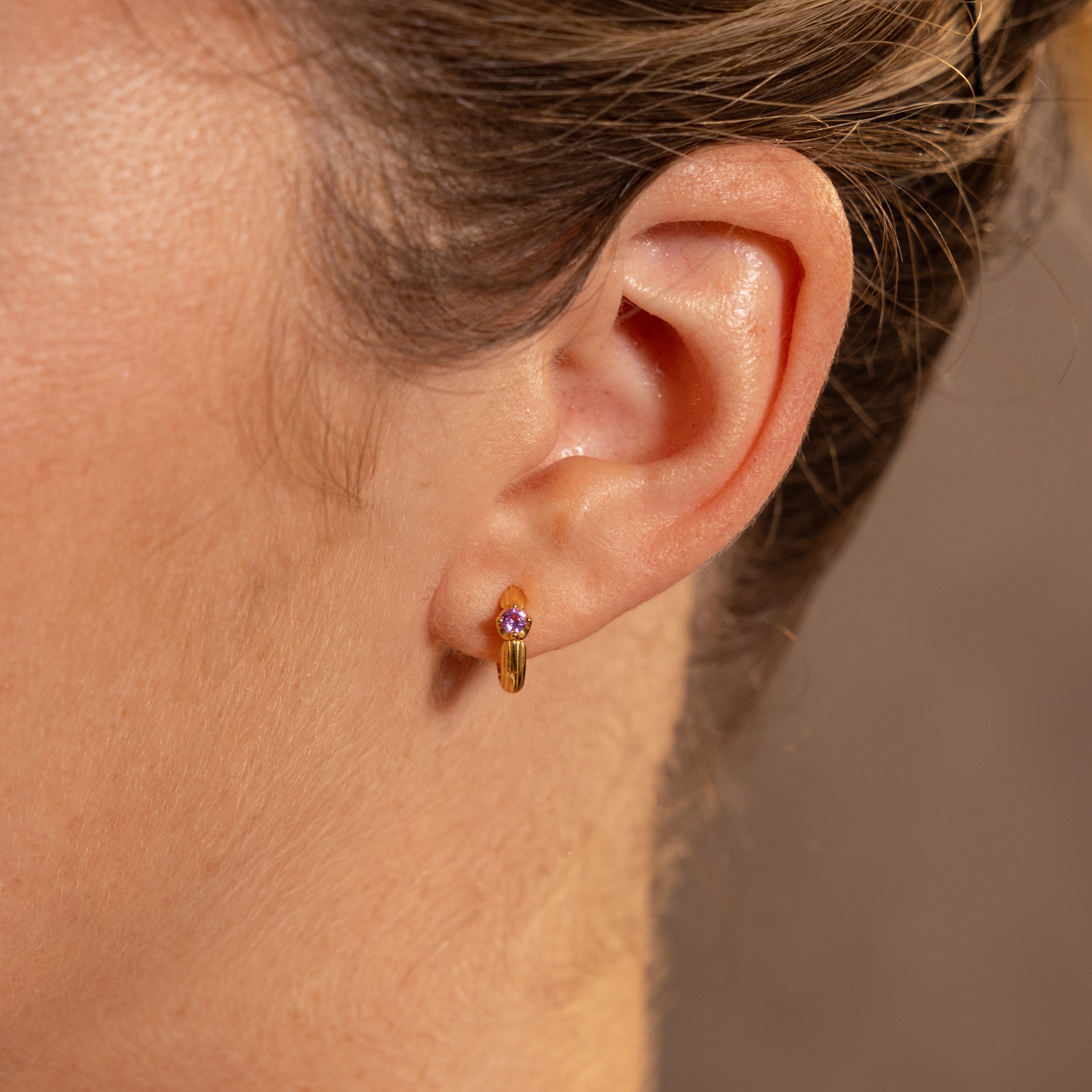 Close-up of a persons ear wearing gold Ribbed Alexandrite Huggies, featuring a pink gemstone for a stylish look.