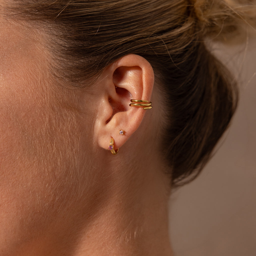 Close-up of a womans ear wearing three gold hoop earrings, including the Ribbed Alexandrite Huggies, an ear cuff, and her hair tied back.