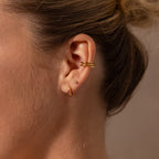 Close-up of a womans ear with Ribbed Emerald Huggies, gold hoop earrings, and an ear cuff—an elegant look and meaningful gift against a neutral background.