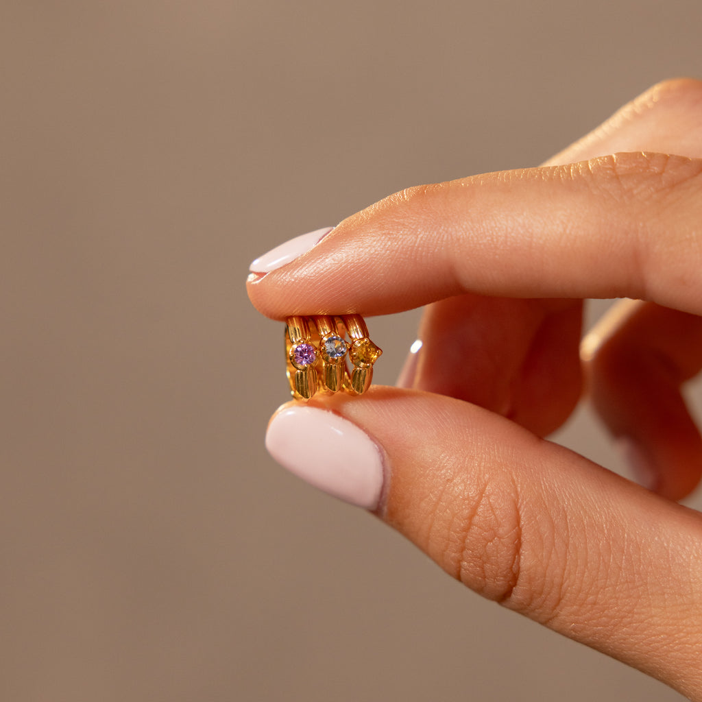 A hand with pale pink nails holds a small gold ring adorned with four colorful gems, inspired by Ribbed Topaz Huggies—a meaningful gift choice.