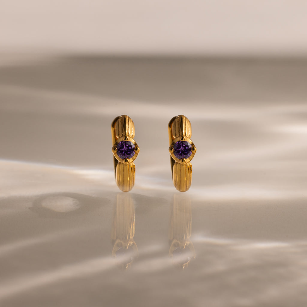 Ribbed Amethyst Huggies—gold hoop earrings with purple gemstones, displayed upright on a marbled surface with soft lighting—perfect for anyone who loves unique birthstone jewelry.