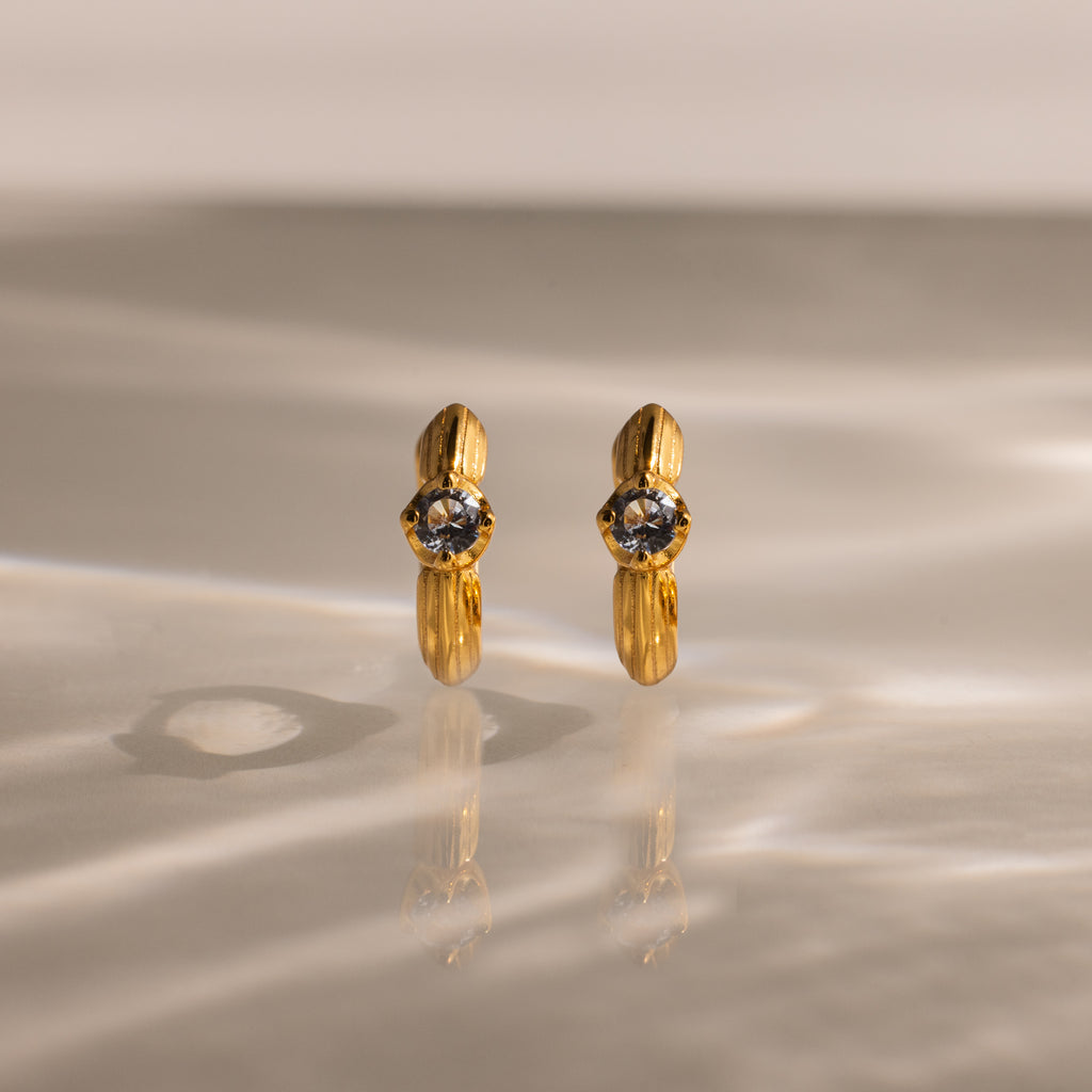 Gold hoop earrings with clear gemstones rest on a reflective surface—these Ribbed Aquamarine Huggies bring personalized elegance to your jewelry collection.