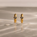 Gold hoop earrings with clear gemstones rest on a reflective surface—these Ribbed Aquamarine Huggies bring personalized elegance to your jewelry collection.