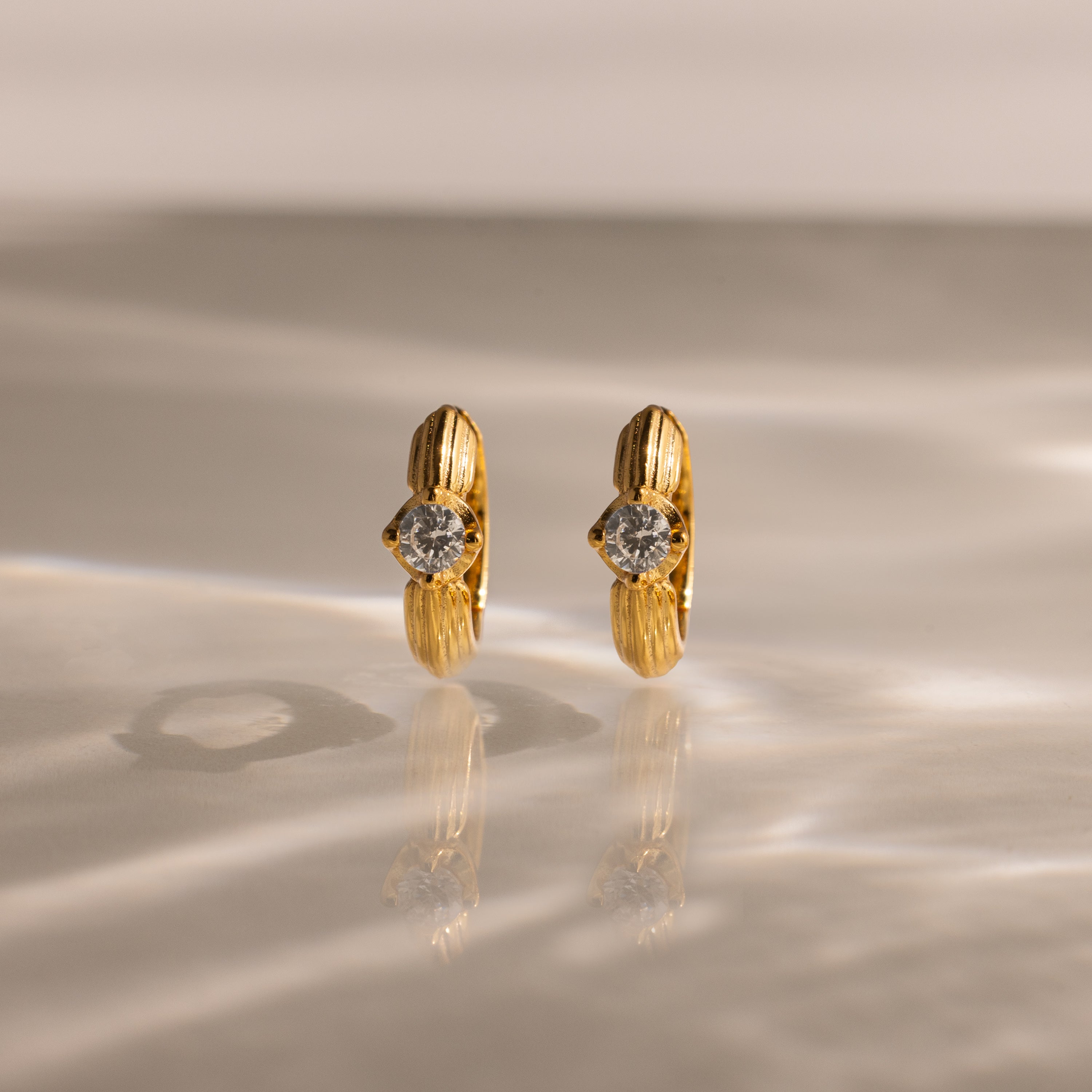 Ribbed Diamond Huggies showcased upright on a reflective, textured surface with soft lighting—an elegant and personalized pair of gold hoop earrings featuring clear gemstones.