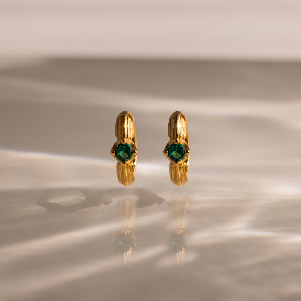 Ribbed Emerald Huggies stand upright on a reflective, softly lit surface—perfect as gold hoop earrings with green gemstones for a meaningful gift.