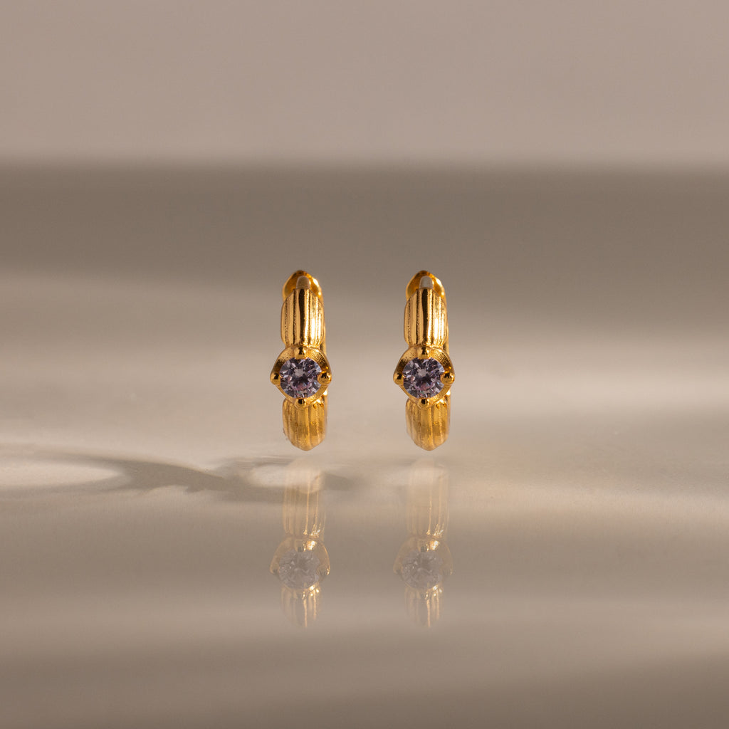 Ribbed Alexandrite Huggies, gold hoop earrings with a small round purple gemstone, stand upright on a reflective surface.