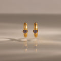 Ribbed Alexandrite Huggies, gold hoop earrings with a small round purple gemstone, stand upright on a reflective surface.
