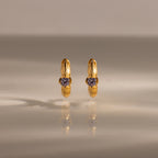 Ribbed Alexandrite Huggies, gold hoop earrings with a small round purple gemstone, stand upright on a reflective surface.