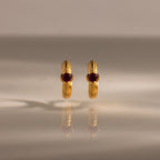 A pair of Ribbed Ruby Huggies with red gemstones stands upright on a reflective surface, illuminated by soft lighting.