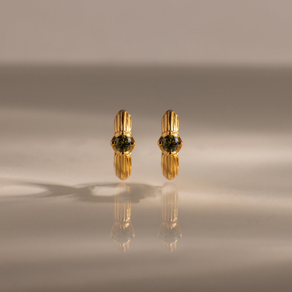 Ribbed Peridot Huggies—gold stud earrings with round green gemstones, shown on a reflective beige surface with soft lighting—ideal for lovers of personalized jewelry.