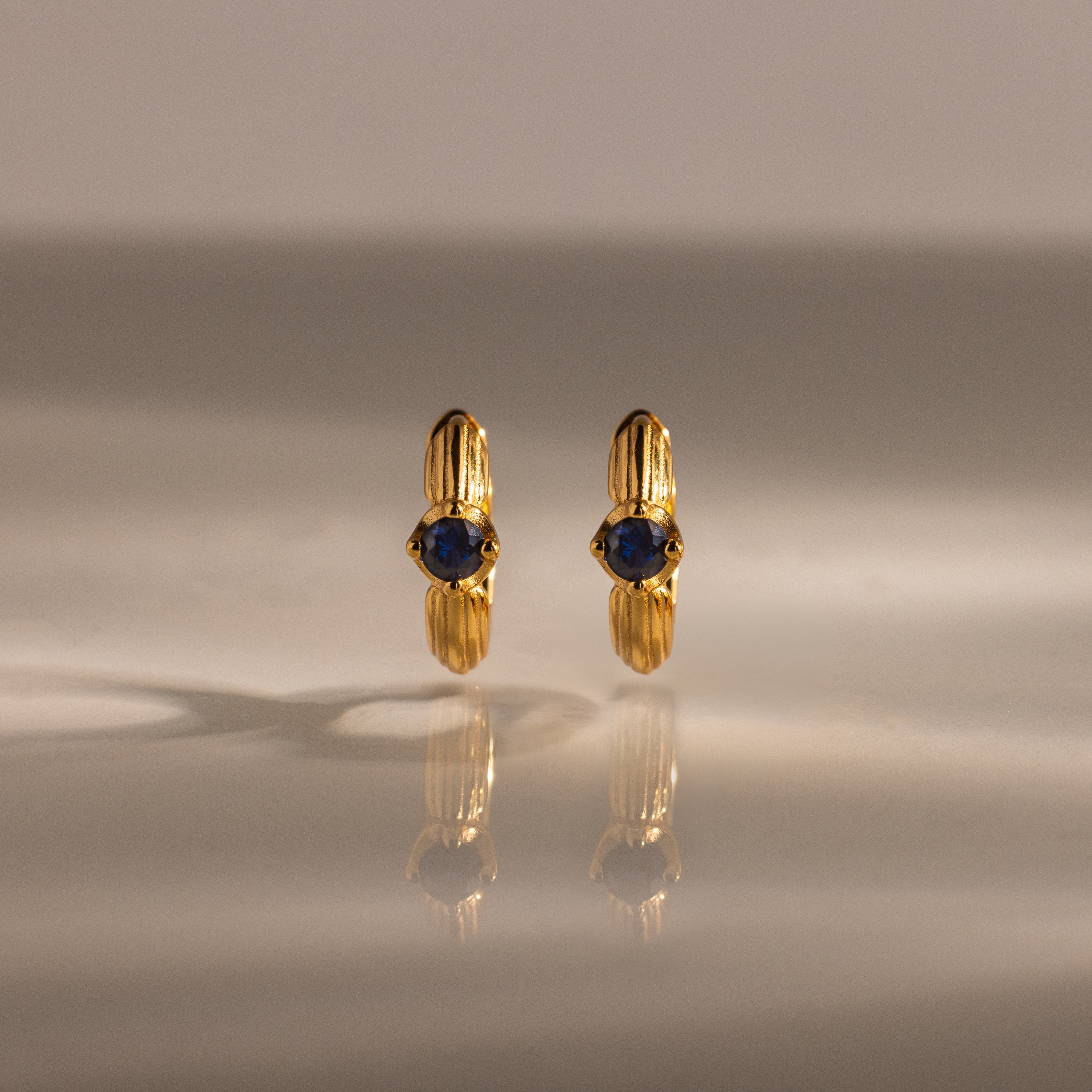 Ribbed Sapphire Huggies—gold stud earrings with blue gemstones—displayed upright on a reflective surface with a soft background, make a meaningful gift for any occasion.
