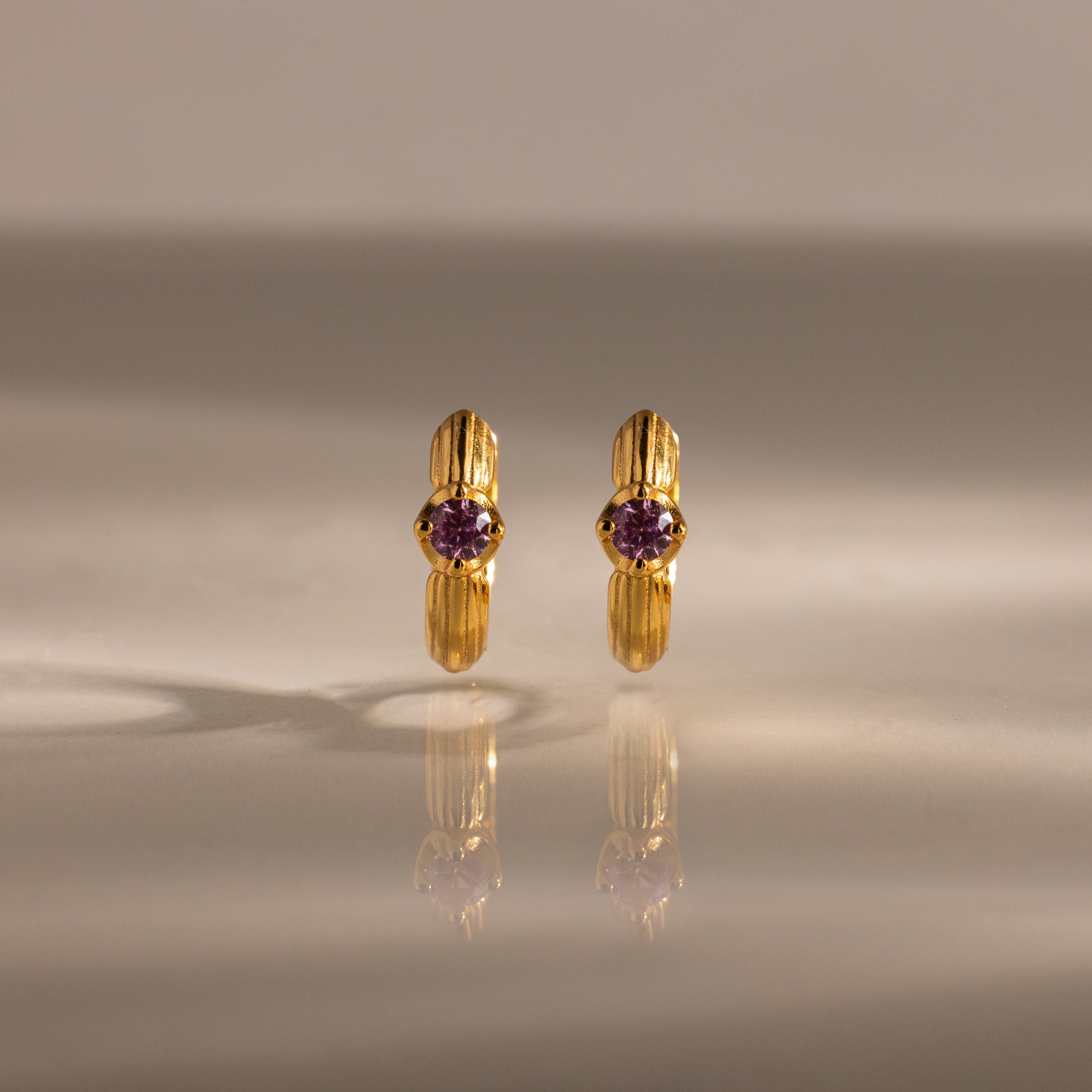 Ribbed Birthstone Huggies are gold hoop earrings with a central purple gemstone, shown upright on a reflective surface—perfect for adding a personalized touch to your jewelry collection.